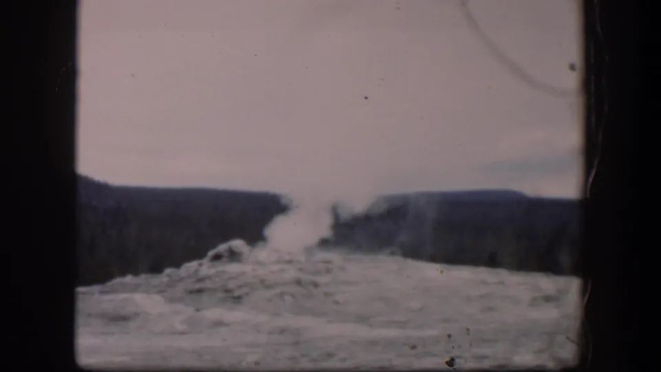 1951 Wyoming 8mm view of a geyser eruption, with steam against a pale sky and dark ridges beyond the vent.