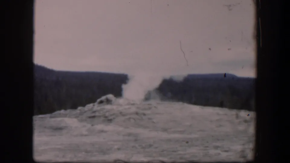 1951 Wyoming home-movie frame with a geyser plume rising from mineral ground, framed by dark hills and visible film wear.