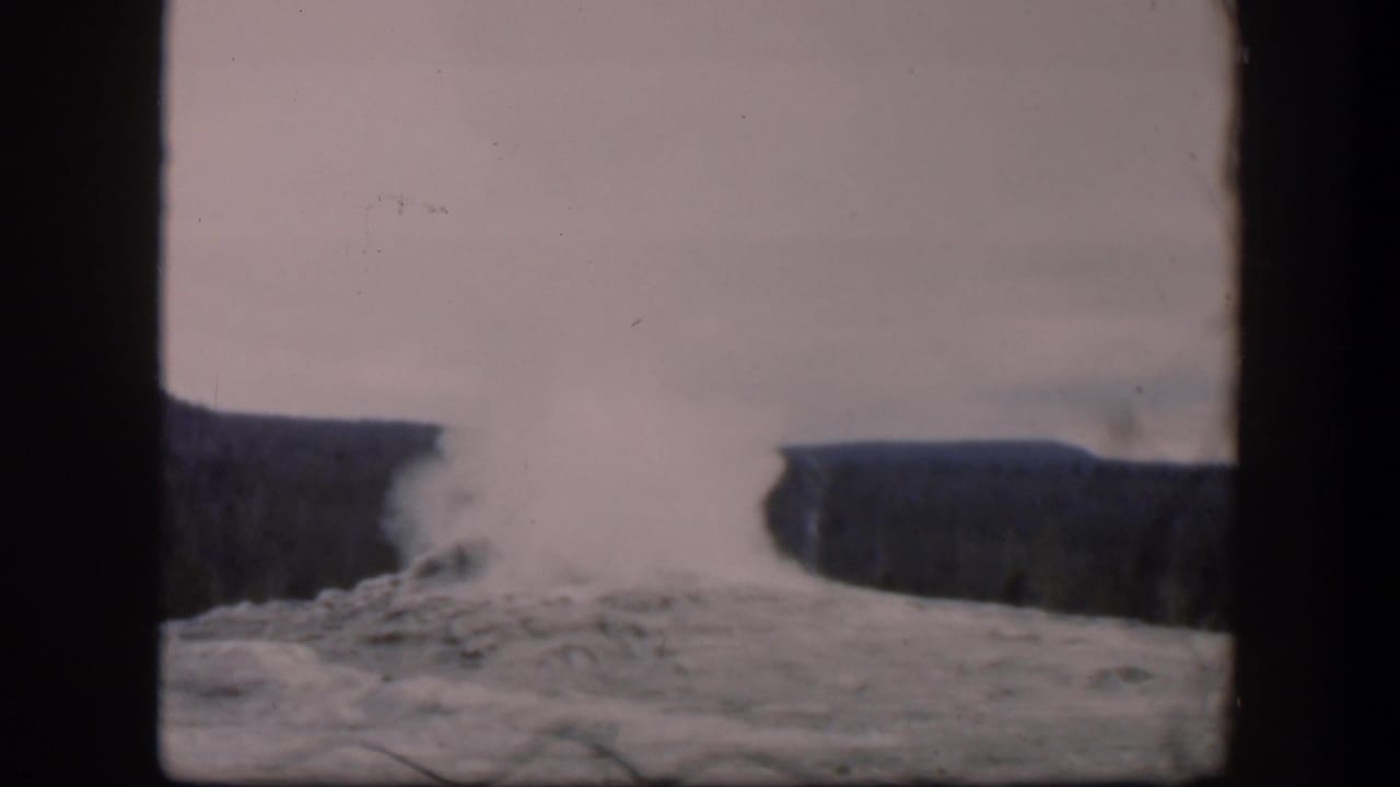 Geyser steam rises from rocky ground with dark hills behind it.