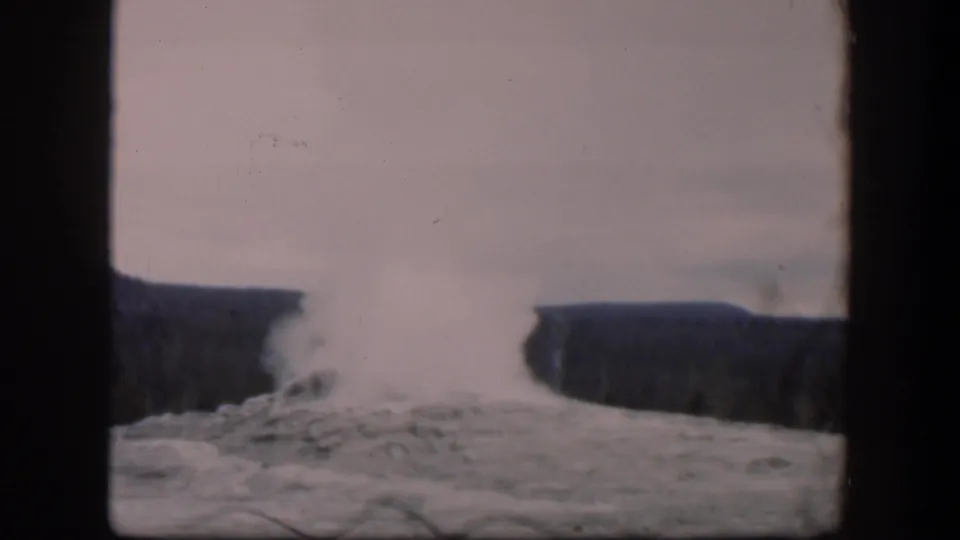 1951 Wyoming 8mm frame showing a geyser plume lifting from pale rocky ground before dark forested ridges.
