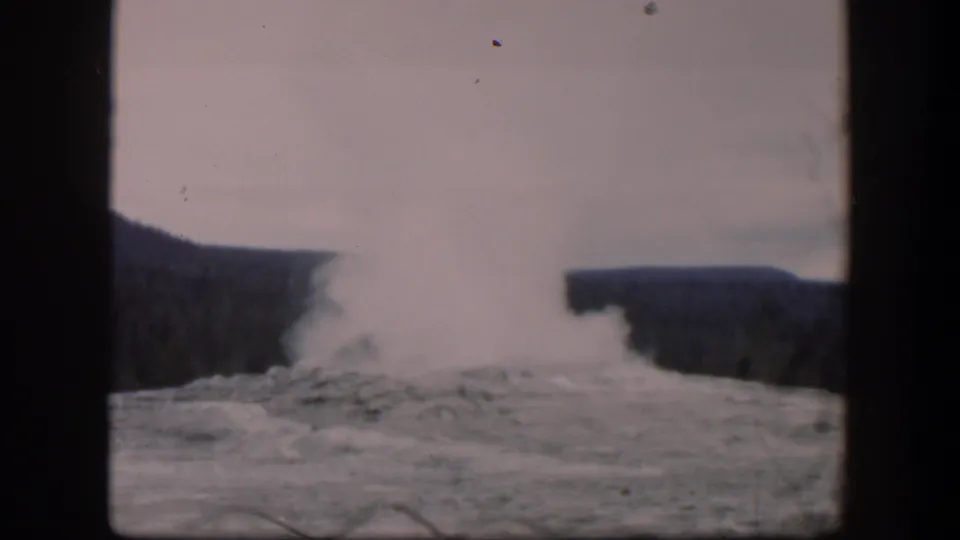 1951 Wyoming home-movie frame with dense geyser steam spreading above rocky mineral ground and distant wooded slopes.