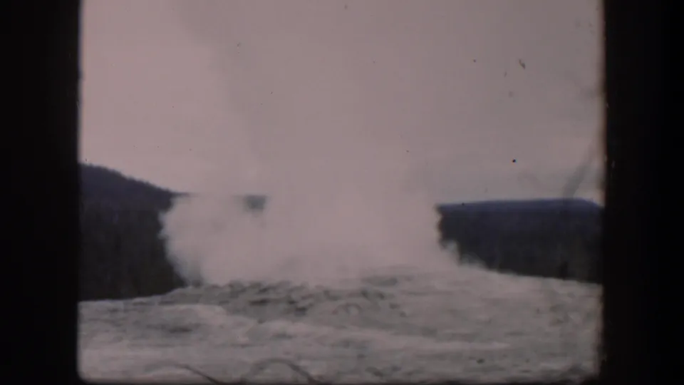 1951 Wyoming frame where the geyser plume lowers and drifts across the rocky basin, backed by dark ridges.