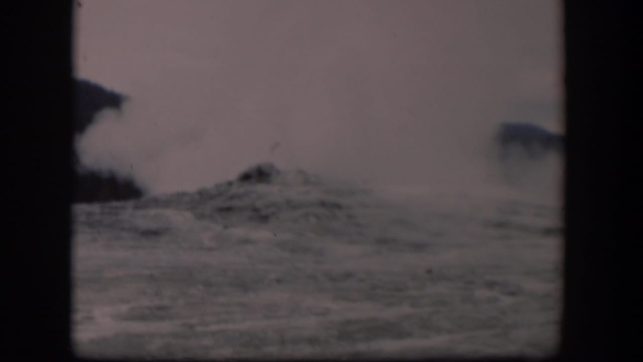 Old Faithful appears mid-eruption in Yellowstone, with steam drifting across a pale sky and rocky ground below.