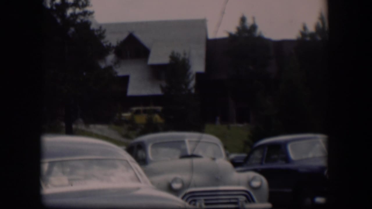 1951 Wyoming home-movie view of parked mid-century cars before trees and a large brown building with a pale sloped roof.