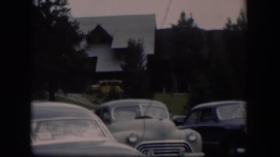 1951 Wyoming home-movie view of parked mid-century cars before trees and a large brown building with a pale sloped roof.
