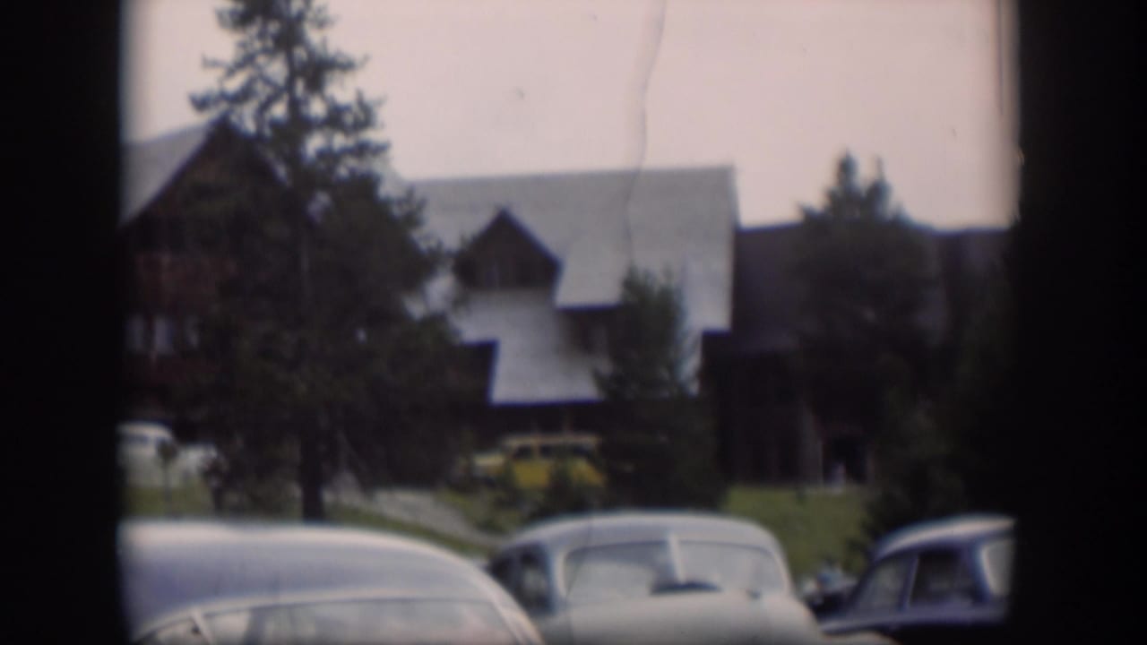 Soft 8mm view in Wyoming of a brown sloped-roof building, tall trees, grass, and several parked mid-century cars.