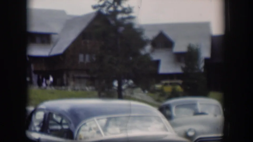 1951 Wyoming 8mm frame showing parked vintage cars in the foreground, a yellow vehicle, trees, and a brown sloped-roof building.