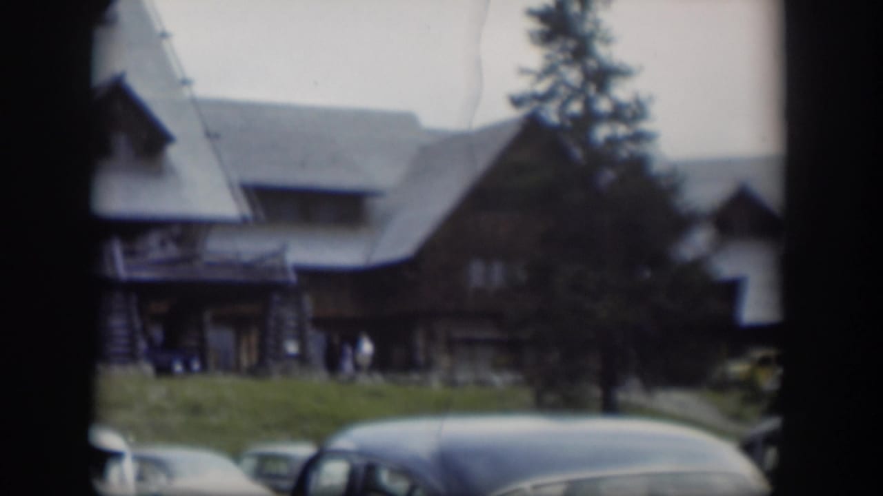 A handheld 8mm view of a Wyoming lodge exterior, with vintage cars in the foreground and visitors gathered near the front porch.