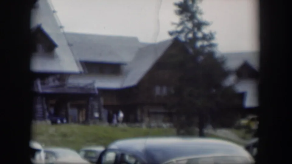 A handheld 8mm view of a Wyoming lodge exterior, with vintage cars in the foreground and visitors gathered near the front porch.