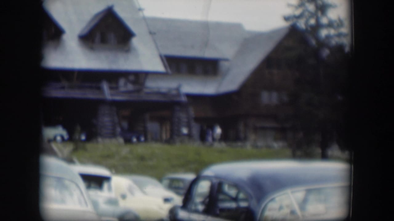 1951 home-movie frame showing the lodge roof and porch above a row of vintage cars on a grassy foreground in Wyoming.