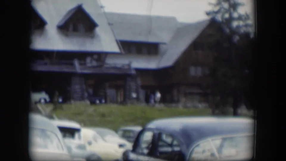 1951 home-movie frame showing the lodge roof and porch above a row of vintage cars on a grassy foreground in Wyoming.