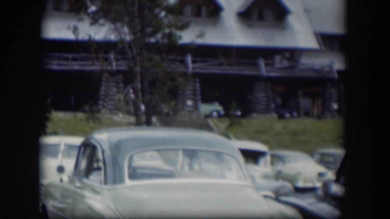 A pale rounded car dominates the foreground outside a log-style building.