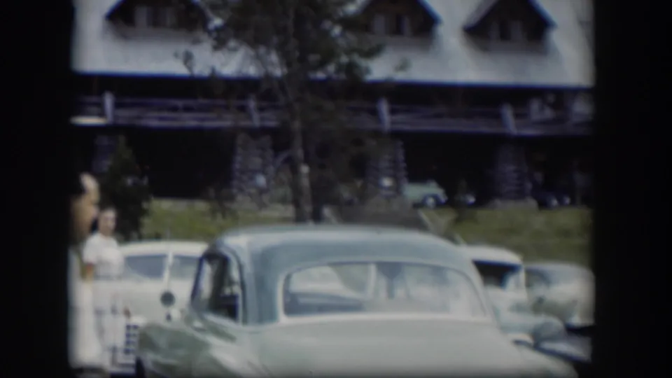 1951 Wyoming home-movie frame showing adults near parked cars outside a log-style gathering building.