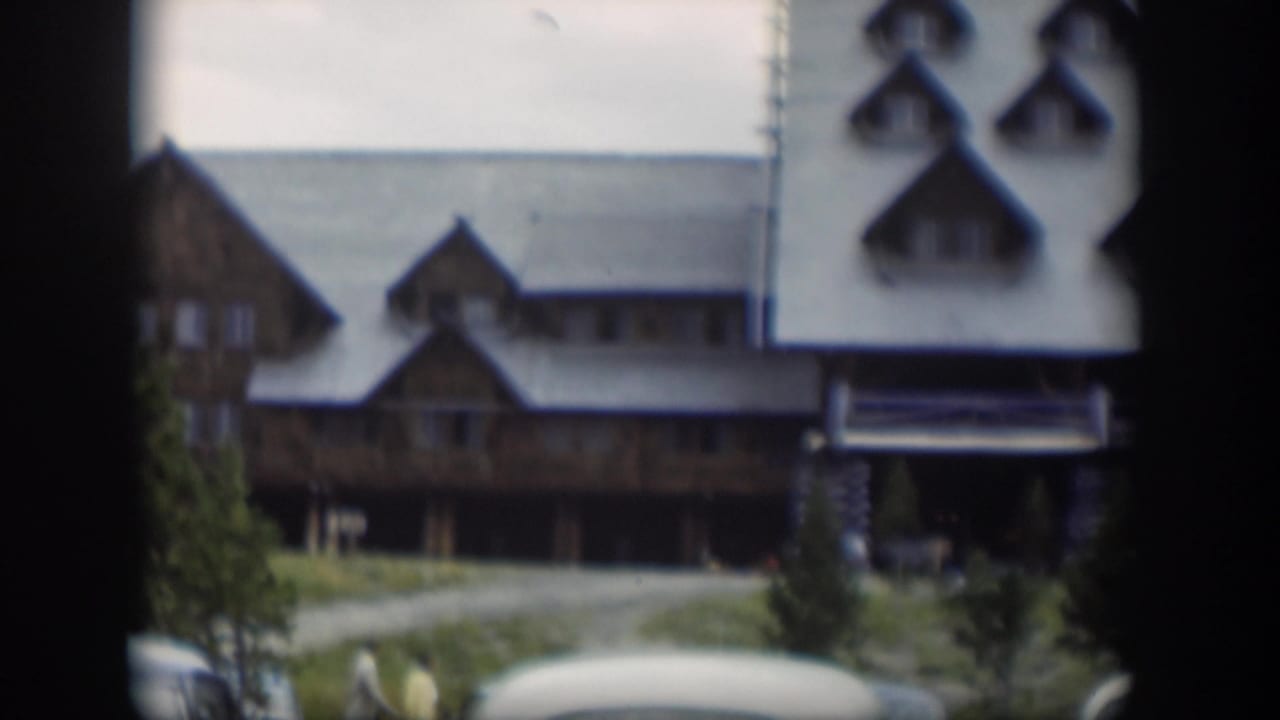 Brown lodge facade with gabled rooflines, trees, driveway, and cars at the edge.