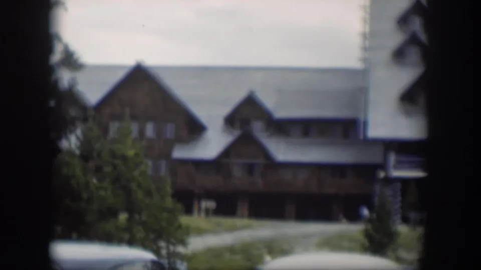Soft 8mm frame of the Wyoming lodge with a gravel approach, green trees, pale roof, and a rounded car partly visible at bottom.