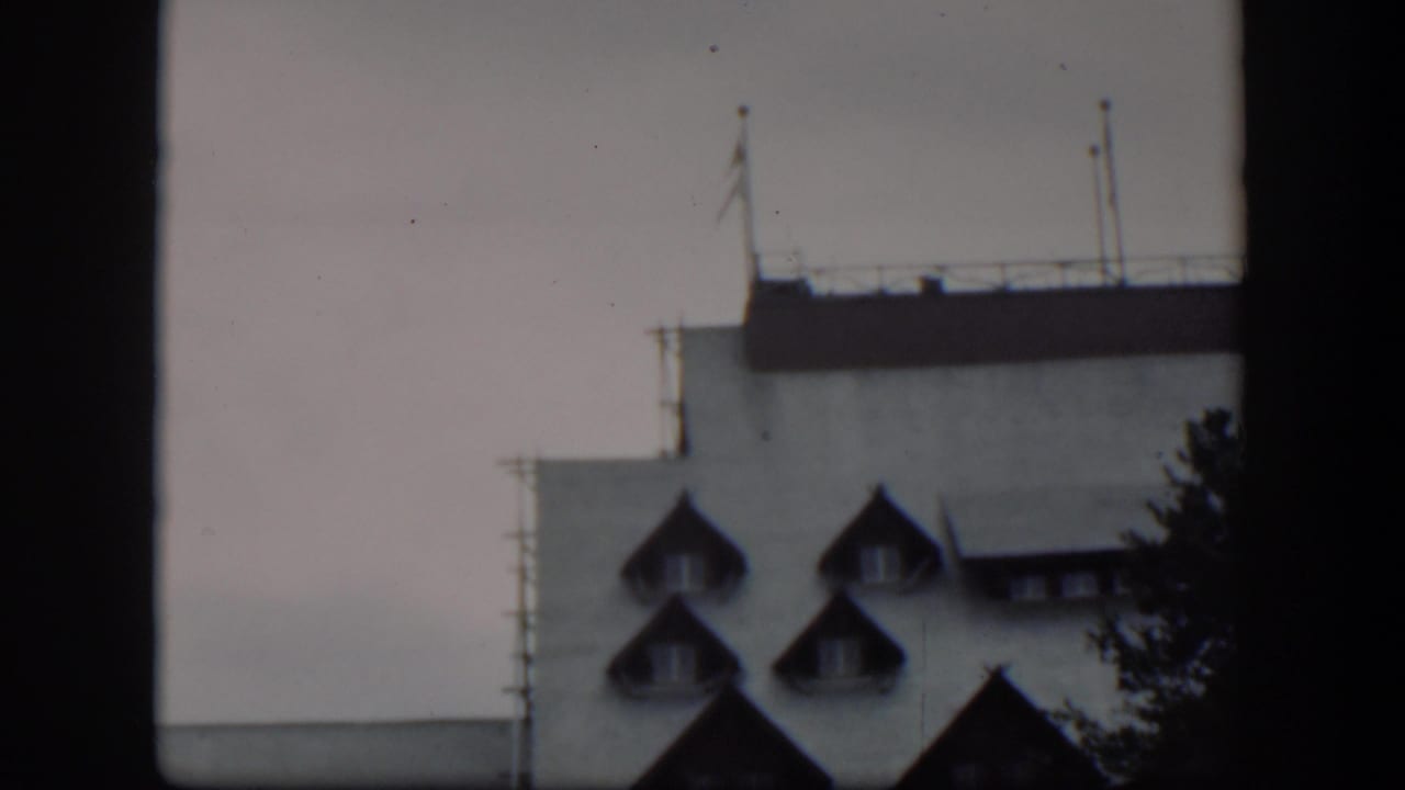 1951 Wyoming frame with the building partly cropped, showing pointed dormers, a flat roof edge, railings, poles, and evergreen branches.