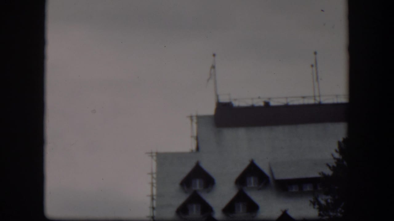 1951 Wyoming 8mm frame showing a pale building roofline, dormer windows, rooftop railings, and flagpoles through a dark window frame.