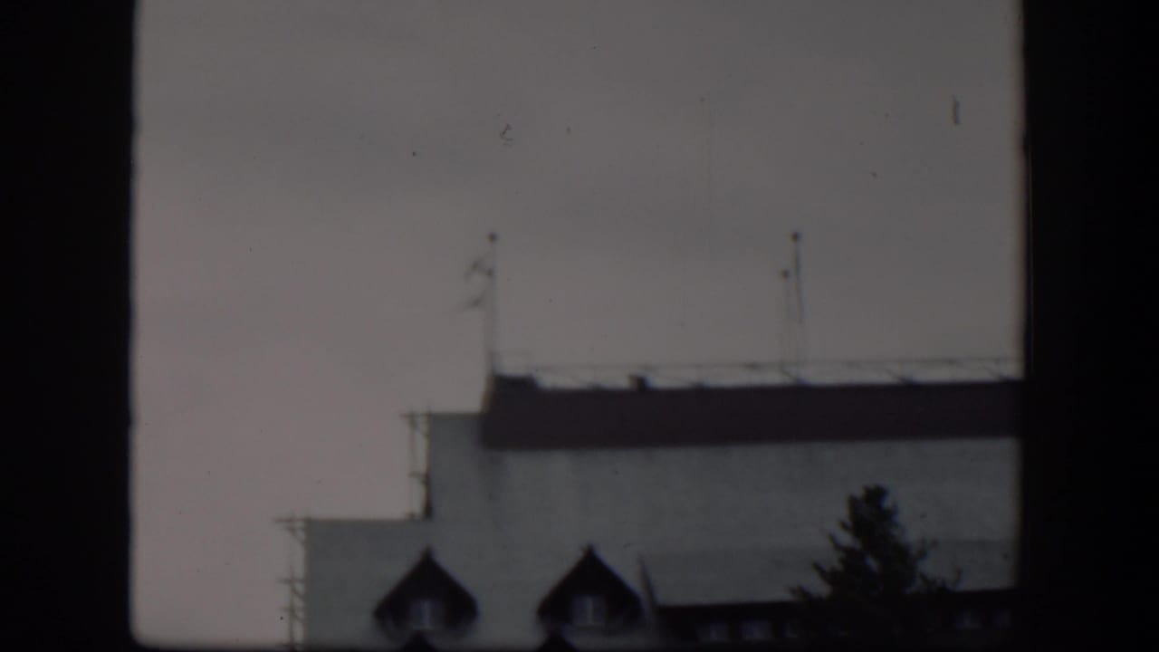 1951 Wyoming 8mm frame with the upper facade of a pale building, dark roof edge, flagpoles, and dormer windows partly obscured by a window frame.