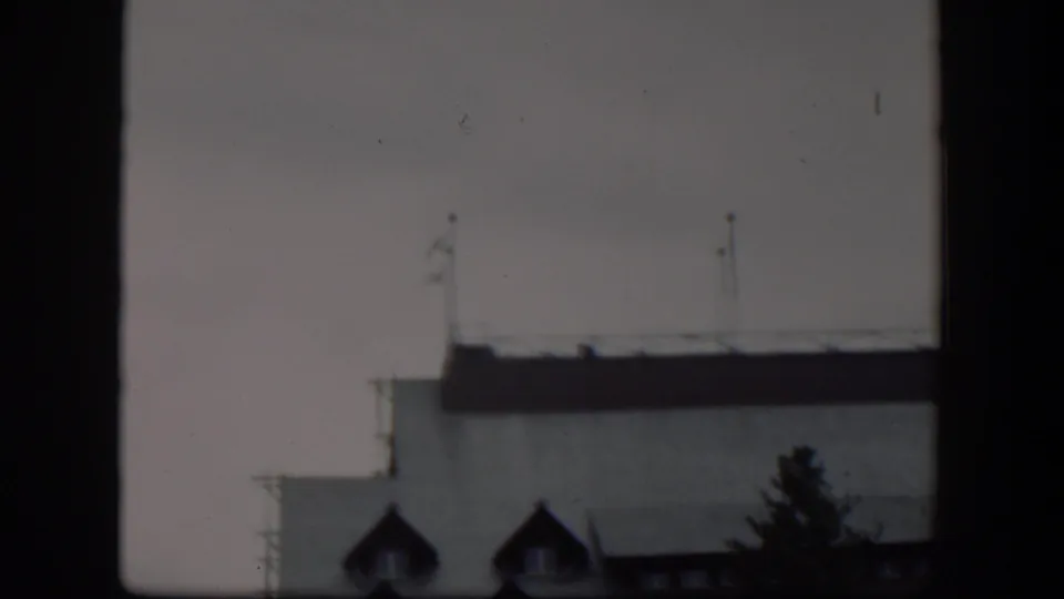 1951 Wyoming 8mm frame with the upper facade of a pale building, dark roof edge, flagpoles, and dormer windows partly obscured by a window frame.