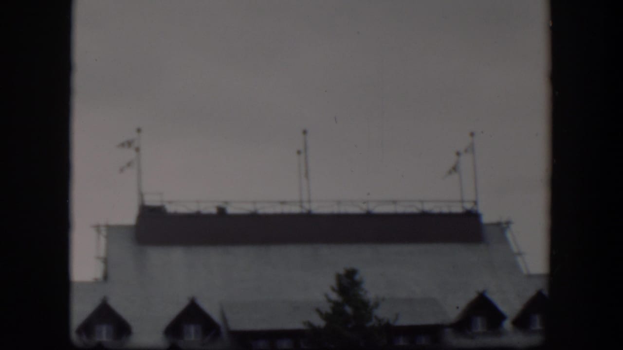 1951 Wyoming 8mm frame showing a closer view of a pale building rooftop with flagpoles, railing, dormer windows, and a tree silhouette.