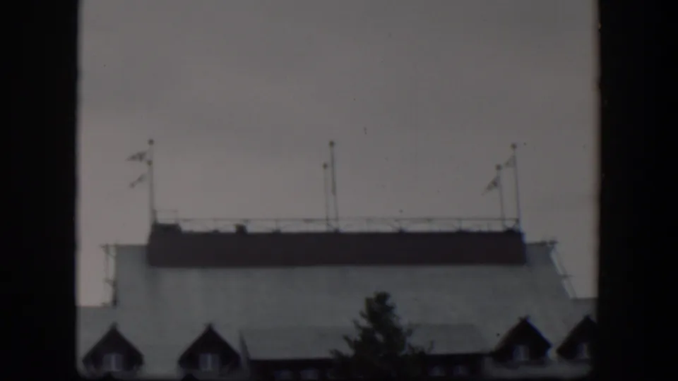1951 Wyoming 8mm frame showing a closer view of a pale building rooftop with flagpoles, railing, dormer windows, and a tree silhouette.