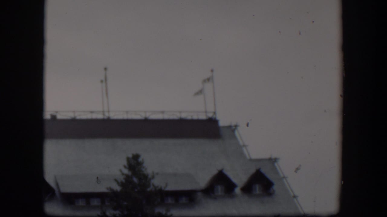 1951 Wyoming 8mm frame of a pale building roofline with rooftop flagpoles, a railing, dormer windows, and a dark tree silhouette in the foreground.