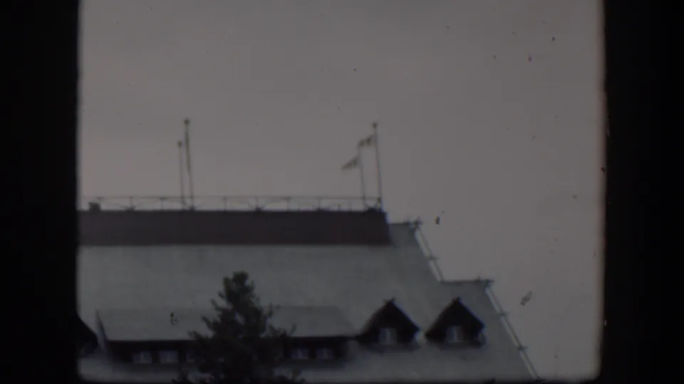 1951 Wyoming 8mm frame of a pale building roofline with rooftop flagpoles, a railing, dormer windows, and a dark tree silhouette in the foreground.