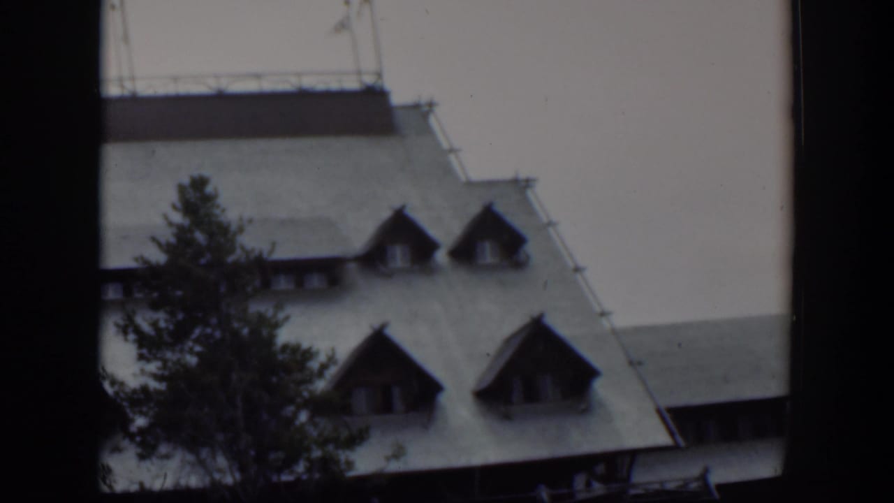 1951 Wyoming home-movie frame with the building roof and dormers seen behind a tall tree and dark gate edge.
