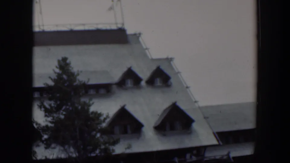 1951 Wyoming home-movie frame with the building roof and dormers seen behind a tall tree and dark gate edge.