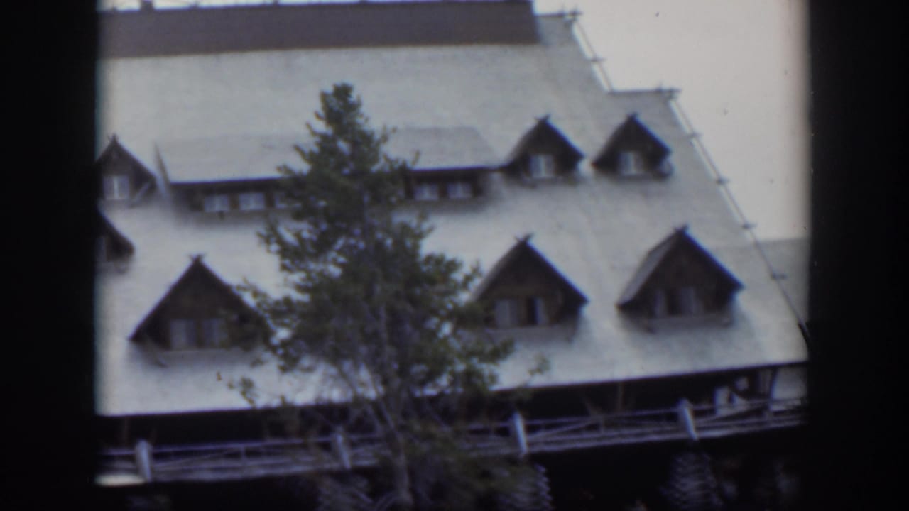 1951 Wyoming frame of a mountain-style lodge facade with dormer windows, porch railing, and evergreen tree.