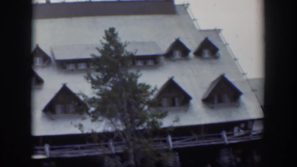 1951 Wyoming frame of a mountain-style lodge facade with dormer windows, porch railing, and evergreen tree.
