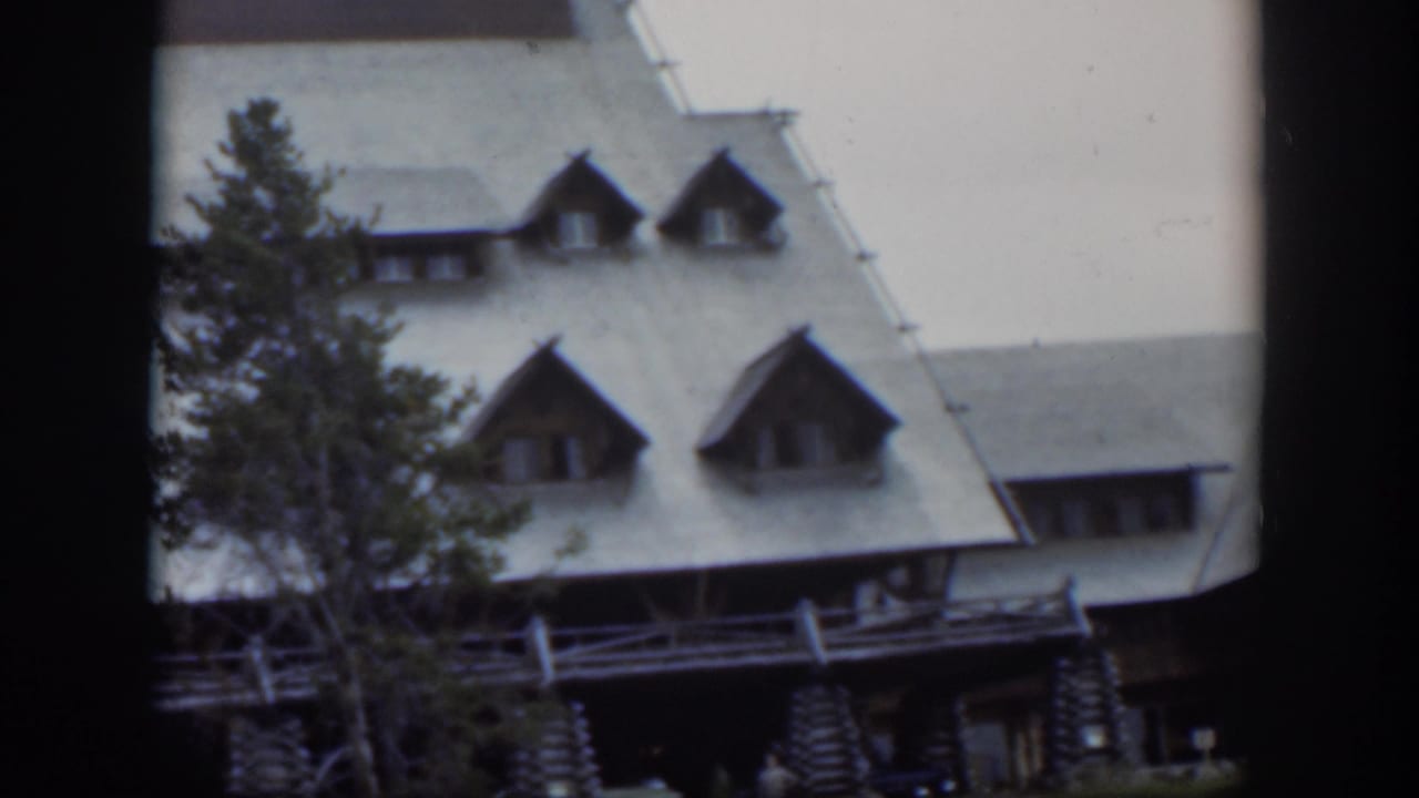 1951 Wyoming lodge view showing dormers, a porch, an evergreen tree, and shaded activity near the entrance.