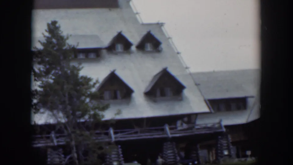1951 Wyoming lodge view showing dormers, a porch, an evergreen tree, and shaded activity near the entrance.