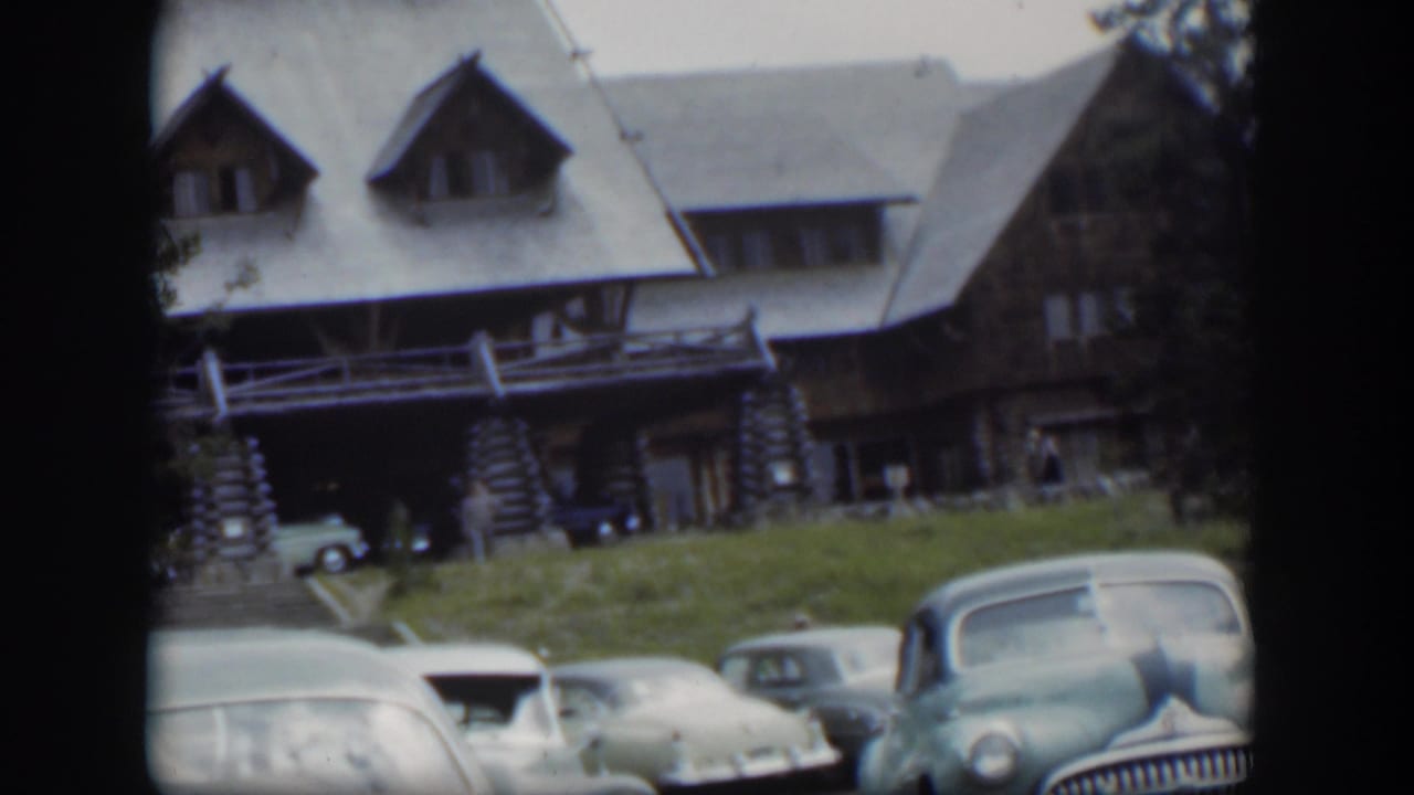 Parked vintage cars below a rustic lodge with stone supports and dormer windows.