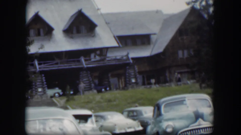 1951 Wyoming home-movie frame showing parked mid-century cars in front of a rustic stone-and-log lodge on a clear day.