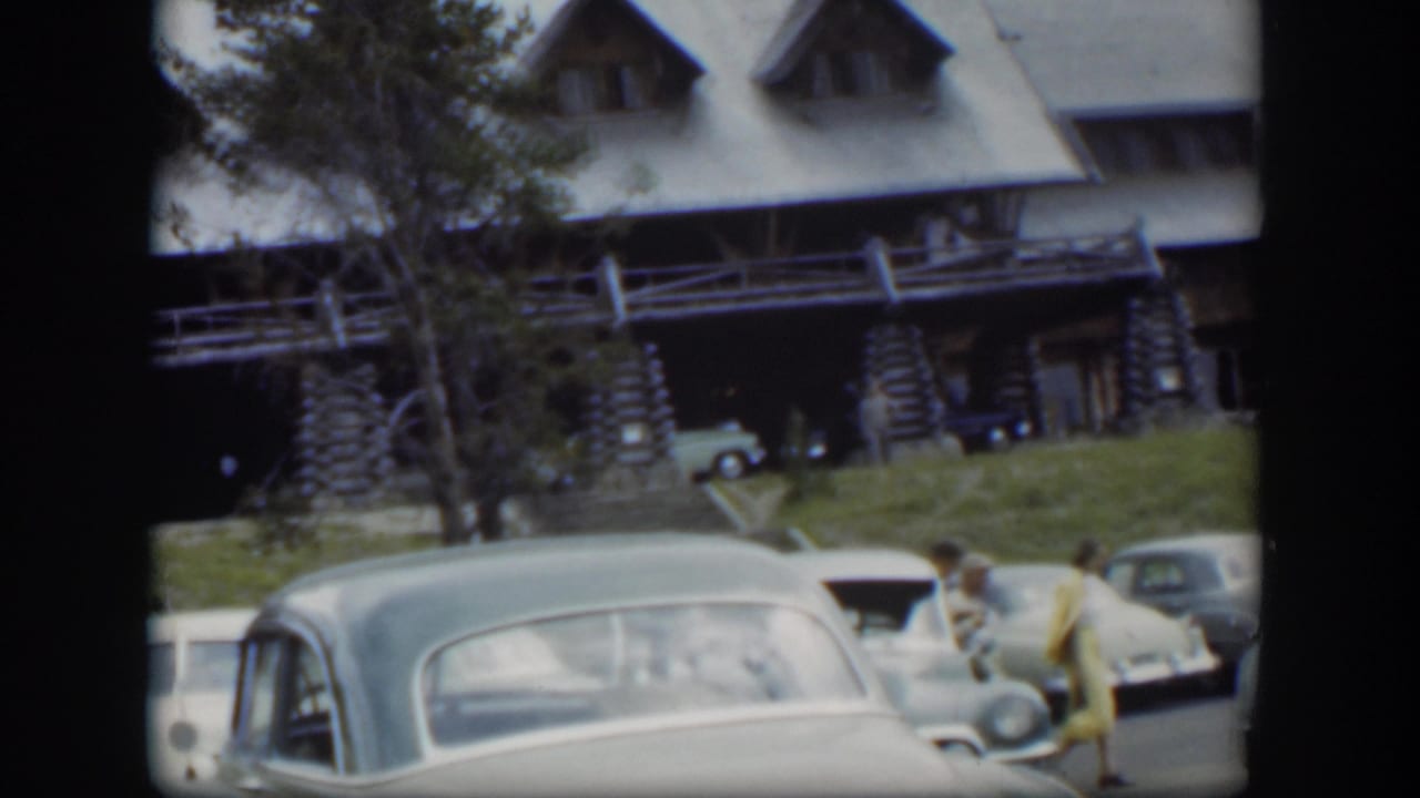 1951 Wyoming frame with a rounded parked car, rustic lodge facade, trees, and a person in yellow walking through the lot.