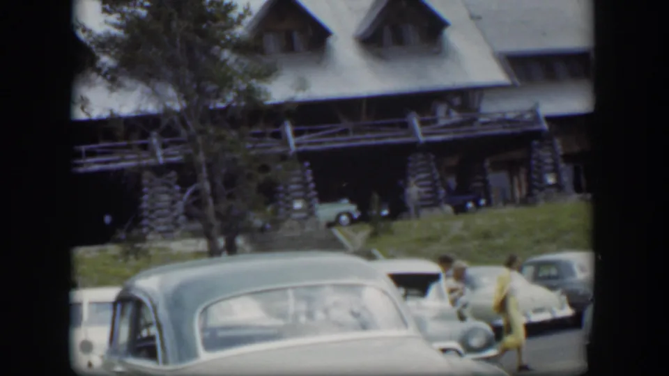 1951 Wyoming frame with a rounded parked car, rustic lodge facade, trees, and a person in yellow walking through the lot.