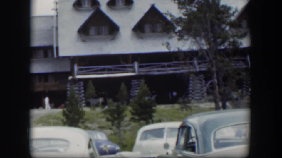 1951 Wyoming frame showing two parked cars below a stone-and-log lodge facade with dormer windows and trees nearby.