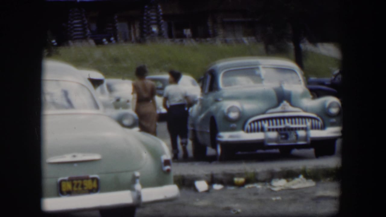 A woman and boy walk away through a 1951 Wyoming parking lot, passing mid-century cars near a rustic lodge.