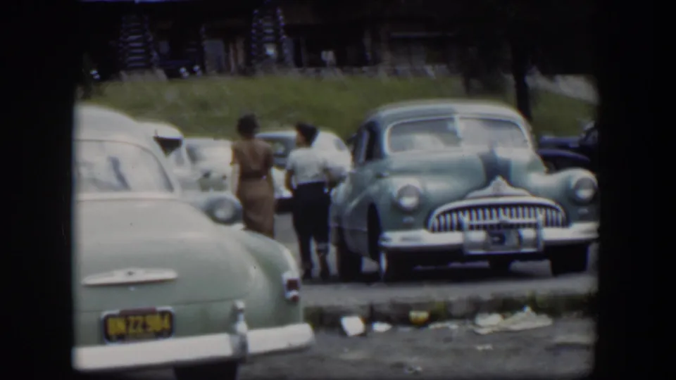 A woman and boy walk away through a 1951 Wyoming parking lot, passing mid-century cars near a rustic lodge.