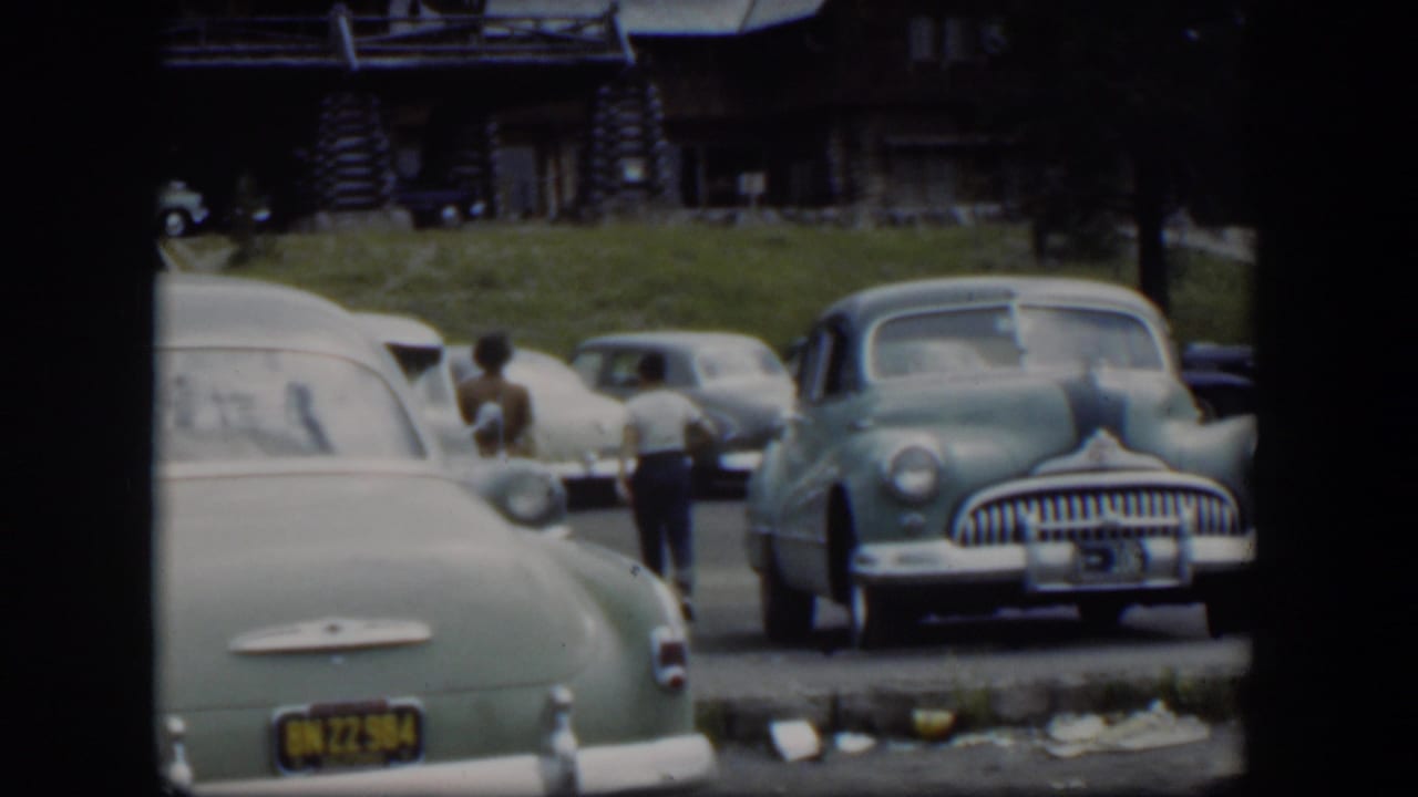 The woman and boy recede among parked cars in a 1951 Wyoming lodge parking lot, with soft 8mm color and handheld framing.