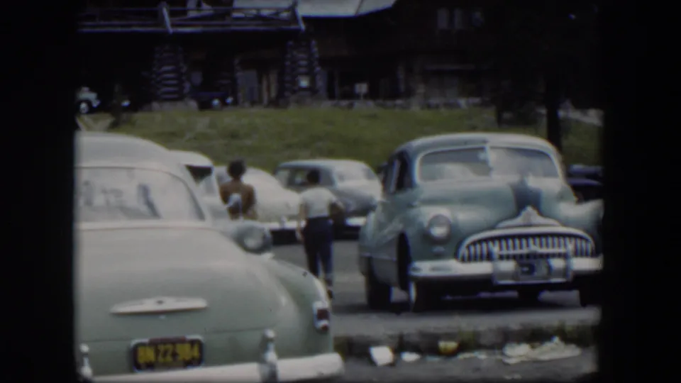 The woman and boy recede among parked cars in a 1951 Wyoming lodge parking lot, with soft 8mm color and handheld framing.