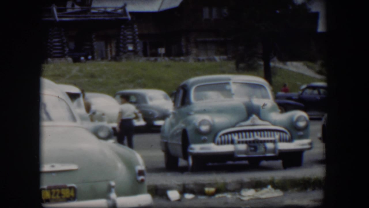 1951 Wyoming 8mm frame showing midcentury cars in a lodge parking lot, including a light green sedan and a man walking nearby.