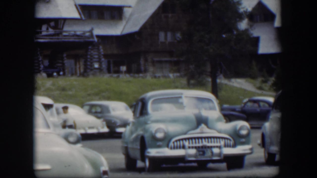 1951 Wyoming 8mm view of a lodge parking area with a light green midcentury sedan, pale cars, and a dark blue car in the background.