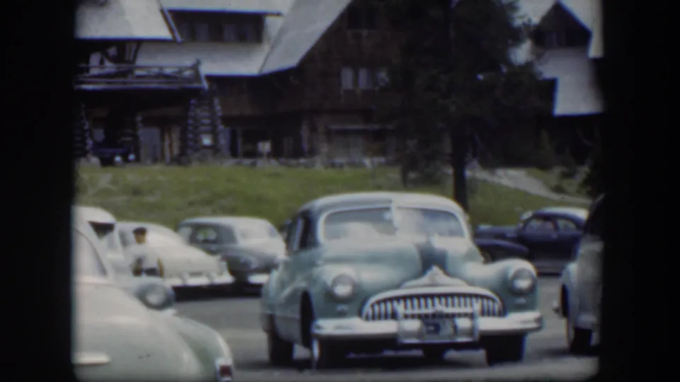1951 Wyoming 8mm view of a lodge parking area with a light green midcentury sedan, pale cars, and a dark blue car in the background.