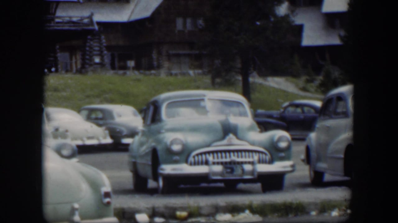 1951 Wyoming home-movie frame with a green sedan centered, other 1950s cars nearby, and a rustic lodge visible behind them.
