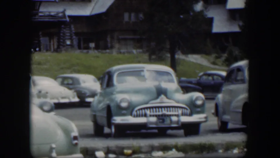 1951 Wyoming home-movie frame with a green sedan centered, other 1950s cars nearby, and a rustic lodge visible behind them.