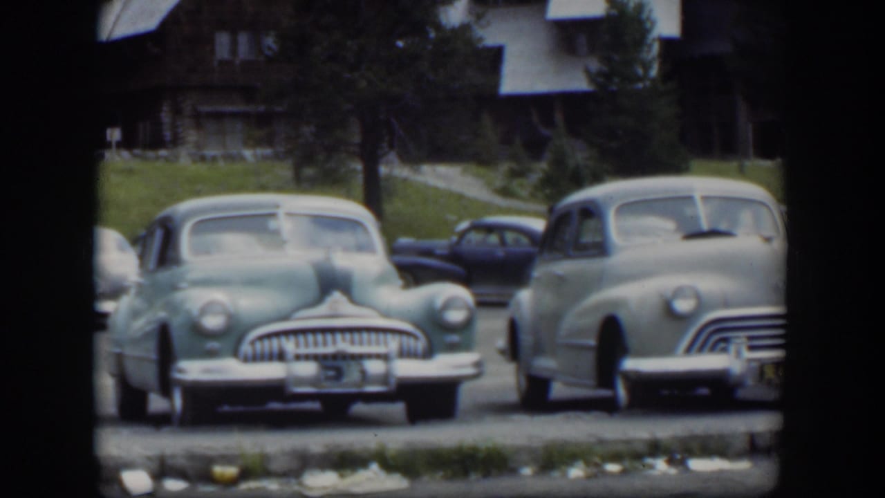1951 Wyoming 8mm frame showing multiple midcentury cars in a lodge lot, with a green sedan centered and a pale car entering the right edge.