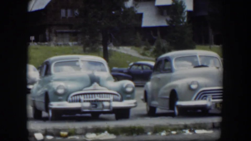 1951 Wyoming 8mm frame showing multiple midcentury cars in a lodge lot, with a green sedan centered and a pale car entering the right edge.