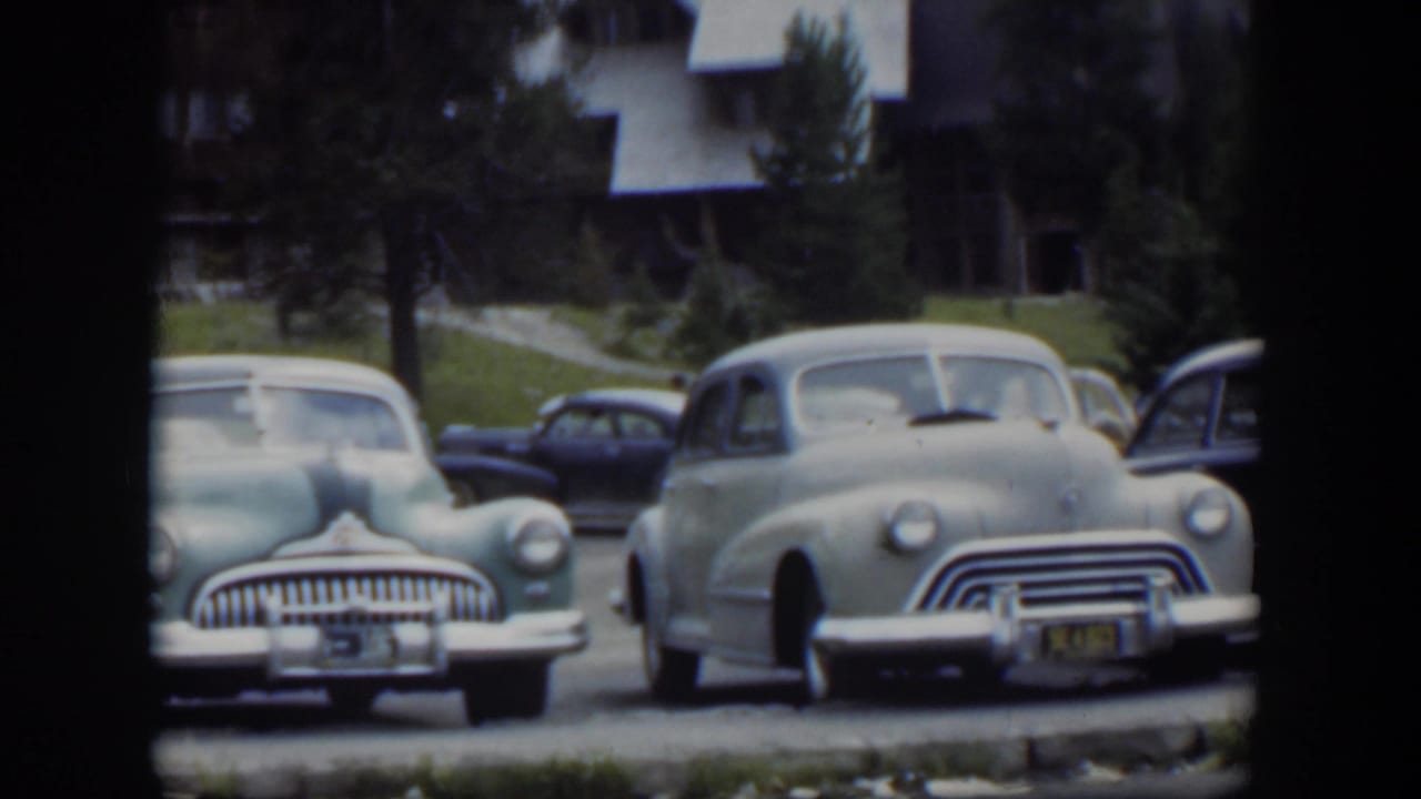 Parked early-1950s cars in front of a lodge-style building and trees.